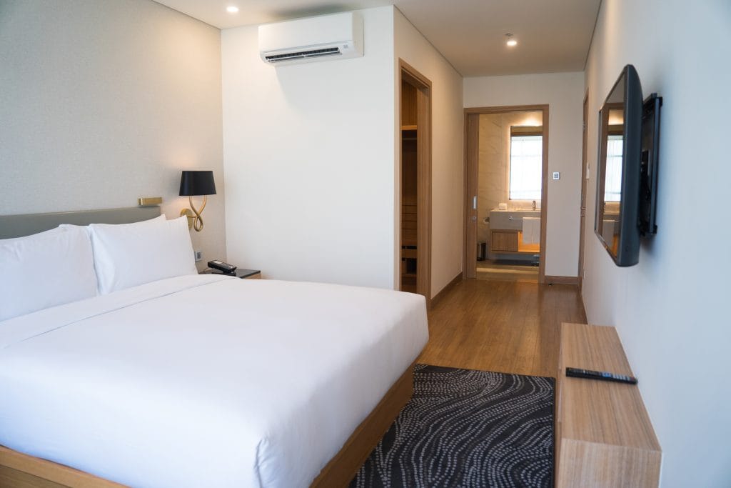 Small hotel room interior with double bed and bathroom. Double bed with white sheets complemented with wall lamp, TV and air conditioner. Hotel room concept