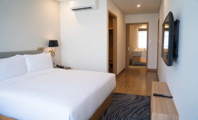 Small hotel room interior with double bed and bathroom. Double bed with white sheets complemented with wall lamp, TV and air conditioner. Hotel room concept